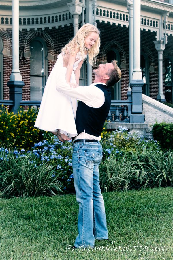 Man lifting young girl in white dress outdoors near brick mansion with flower gardens.