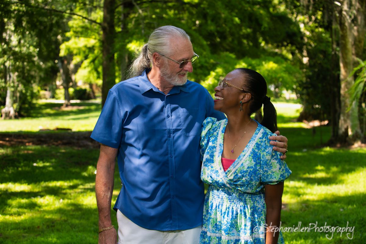 An interracial couple smiling at each other in a lush green park, man in blue shirt, woman in floral dress.