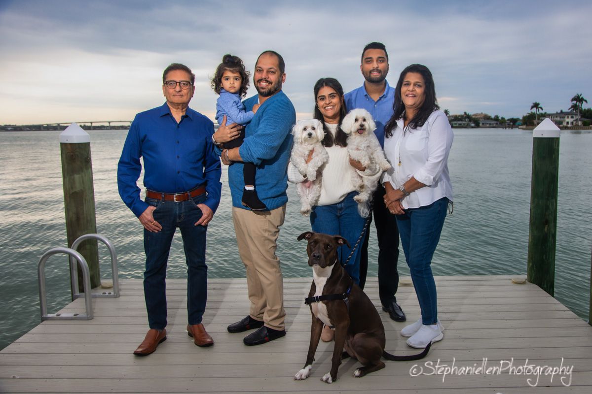 Extended family group with two dogs poses on a waterfront dock at dusk for a professional portrait.
