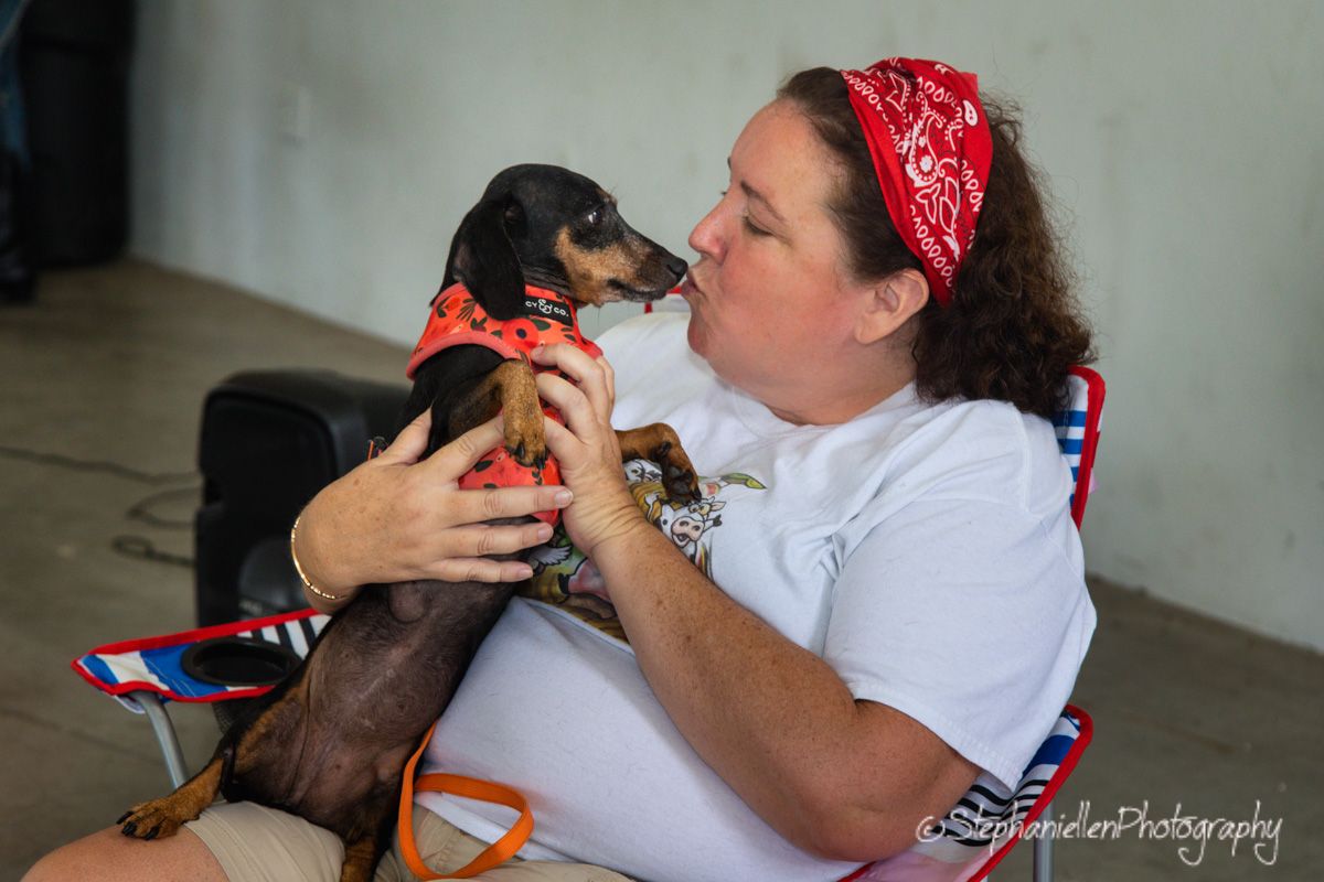 Woman in white shirt and red bandana lovingly kisses her dachshund dog dressed in matching red bandana outdoors.