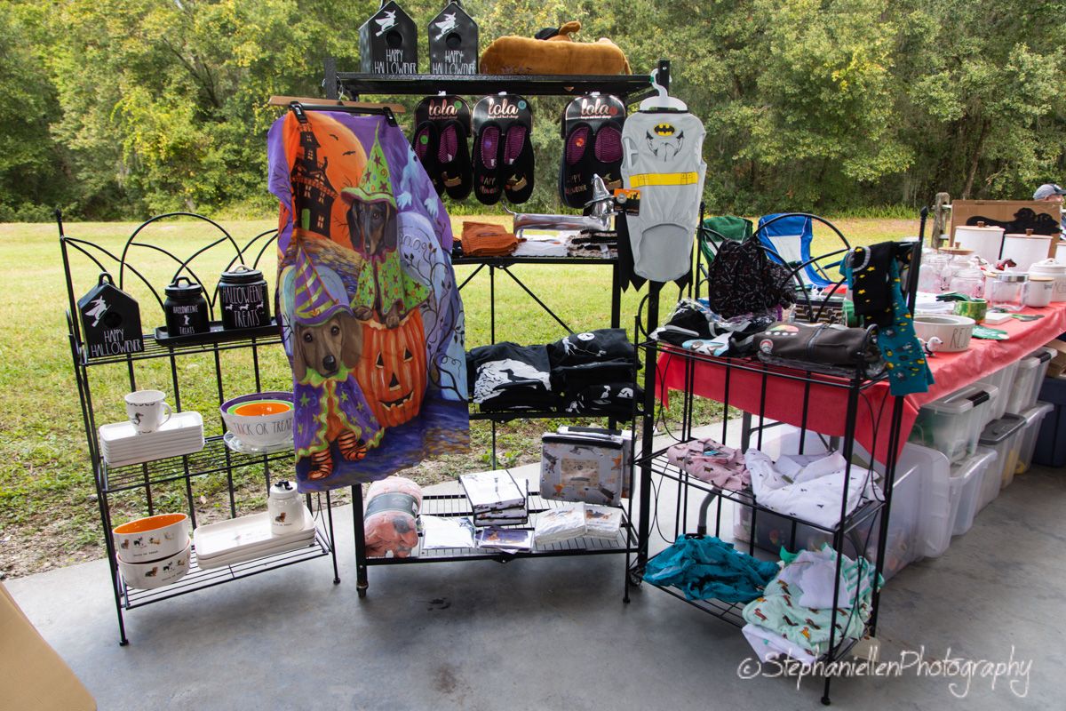 Outdoor garage sale display with black metal shelves holding bags, accessories, and Halloween decor on a paved surface.