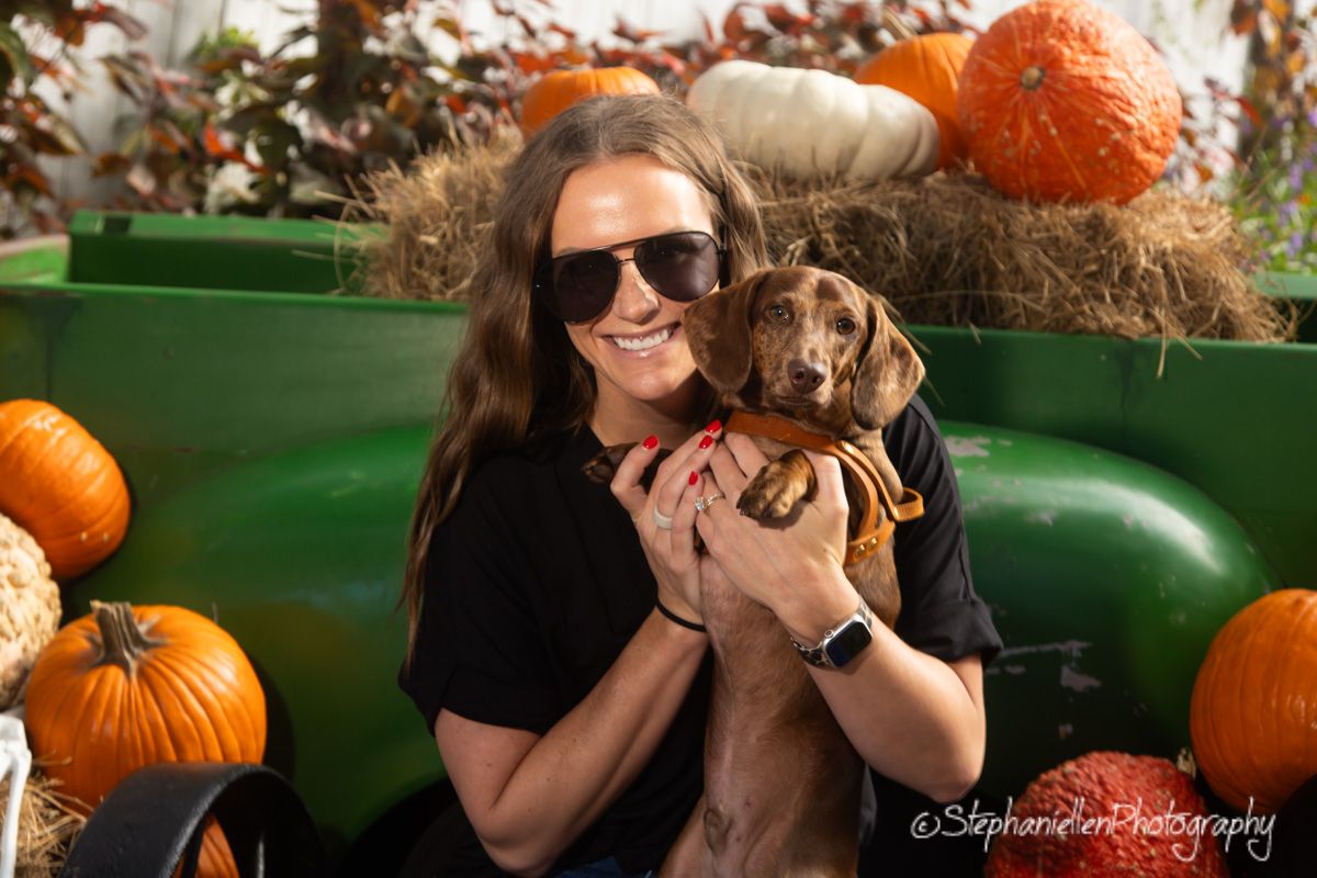 Smiling woman in sunglasses holding a dachshund at a festive fall pumpkin patch display.