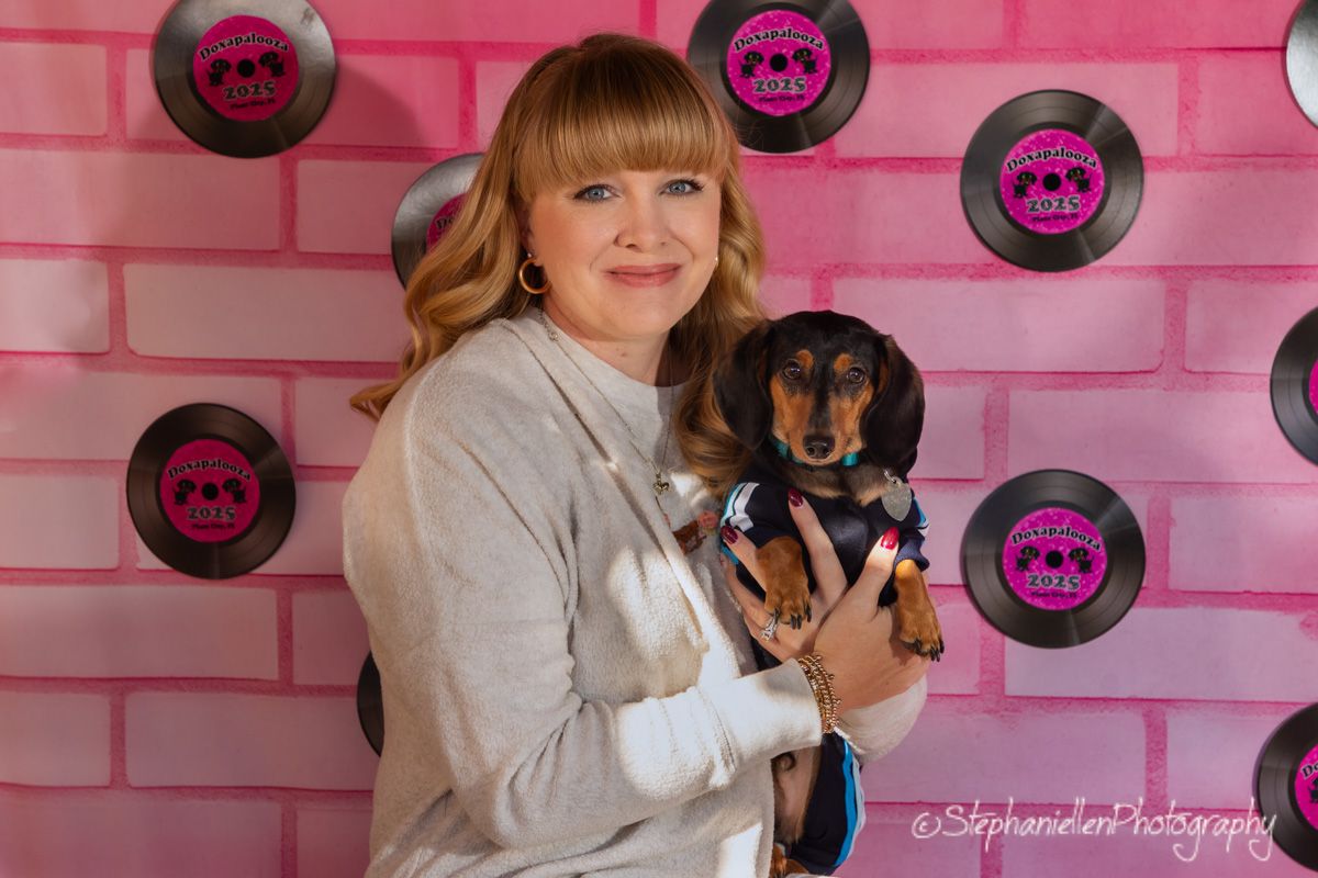Woman holding dressed dachshund dog in front of pink vinyl record backdrop.