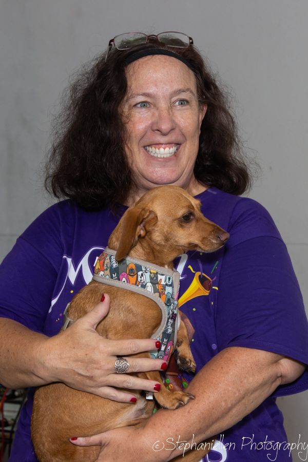 Smiling woman in purple shirt holding a small brown dachshund wearing a colorful bandana at an indoor event.