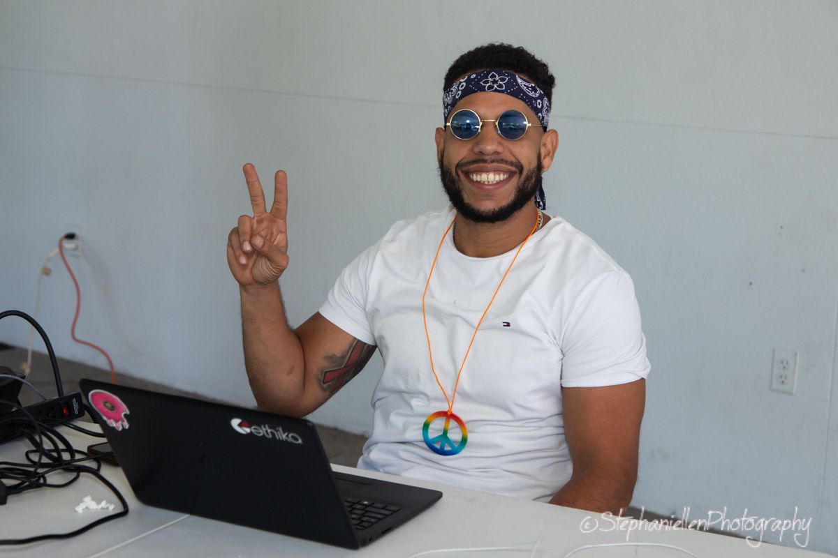 Smiling man wearing peace sign necklace and bandana gives peace sign while sitting at laptop outdoors.