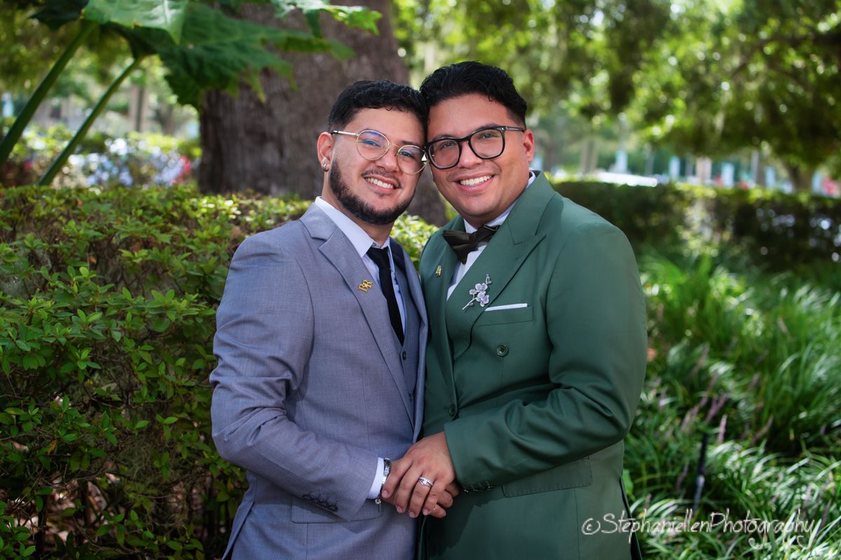Two grooms in gray and green suits smiling together at outdoor wedding.