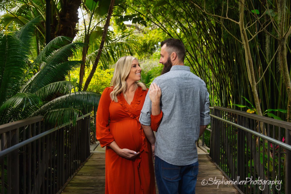 Pregnant woman in red dress and bearded man sharing a tender moment on a bamboo bridge.
