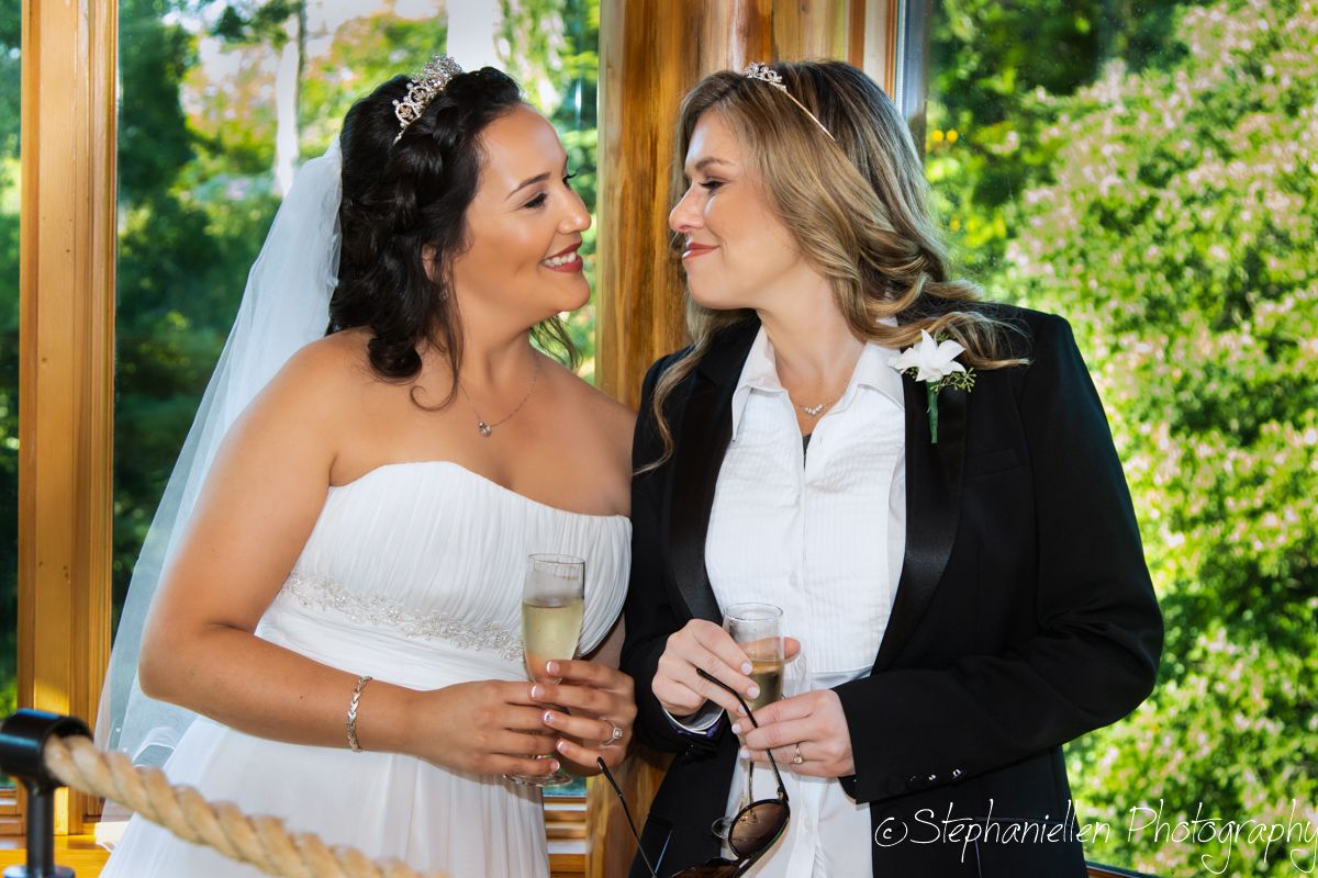 Two brides smiling and toasting with champagne glasses in an elegant indoor ceremony venue.