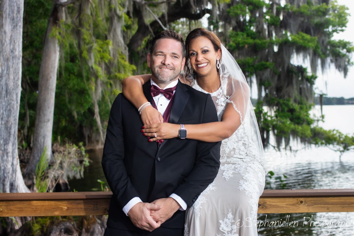 Smiling bride in lace gown and groom in dark suit with burgundy bow tie pose together near lakeside dock.