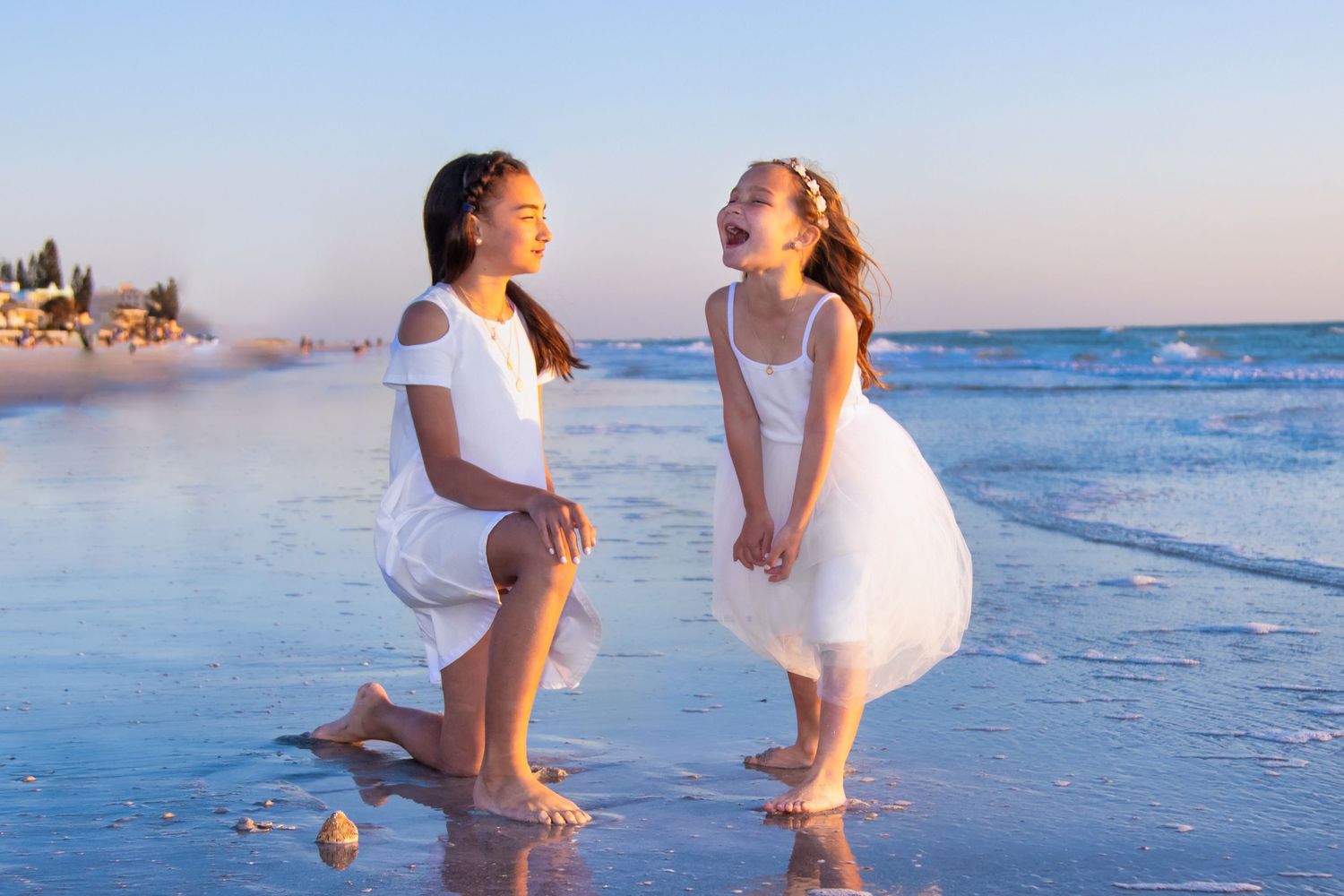 Two young girls in white dresses laugh and play together on a sunny beach with gentle waves.