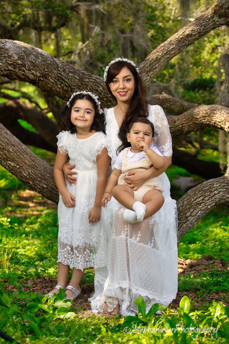 A mother in white lace dress holds baby while daughter in white dress poses outdoors among mossy oak trees.