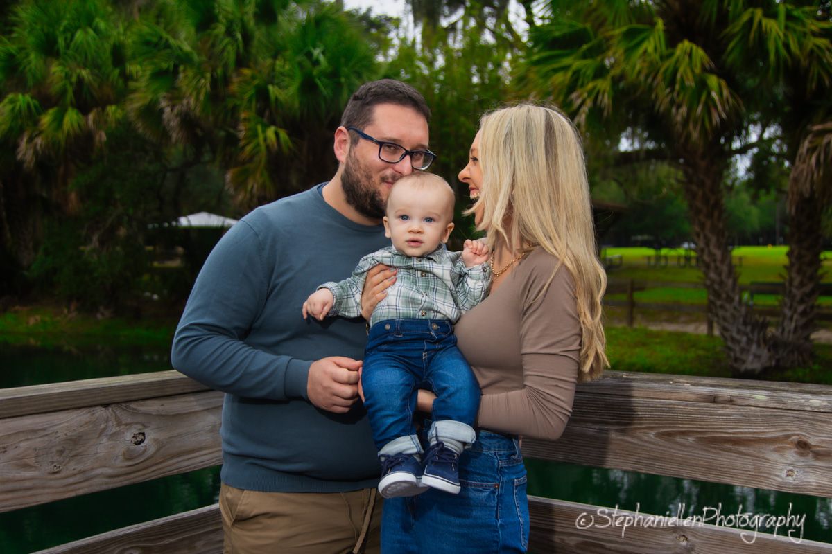 A young family with a baby poses outdoors near lush green palm trees in a warm, candid portrait session.