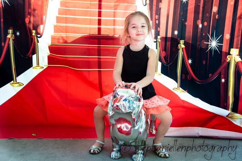 Young girl in black dress posing on silver bulldog statue on red carpet with gold stanchions backdrop.