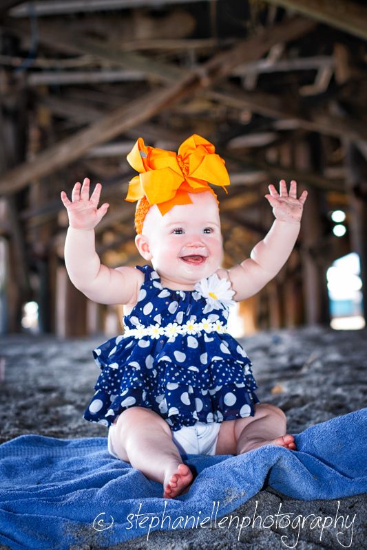Happy baby girl in navy polka dot dress with orange bow headband raising hands under pier.