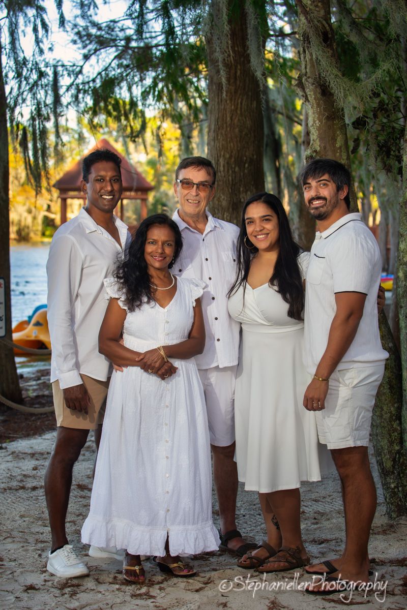 Family of five posing together in white outfits outdoors near a lake surrounded by trees.