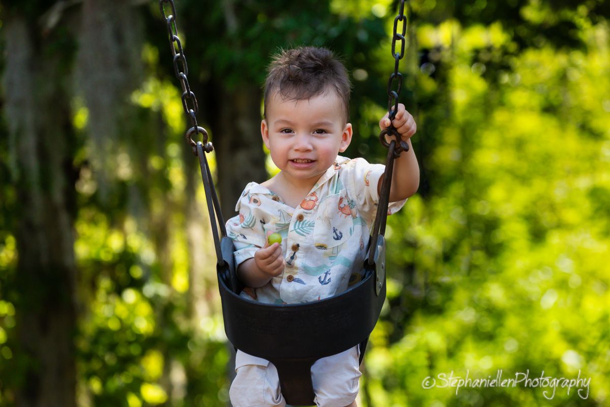 Smiling toddler boy sitting in a black bucket swing outdoors surrounded by lush green trees.