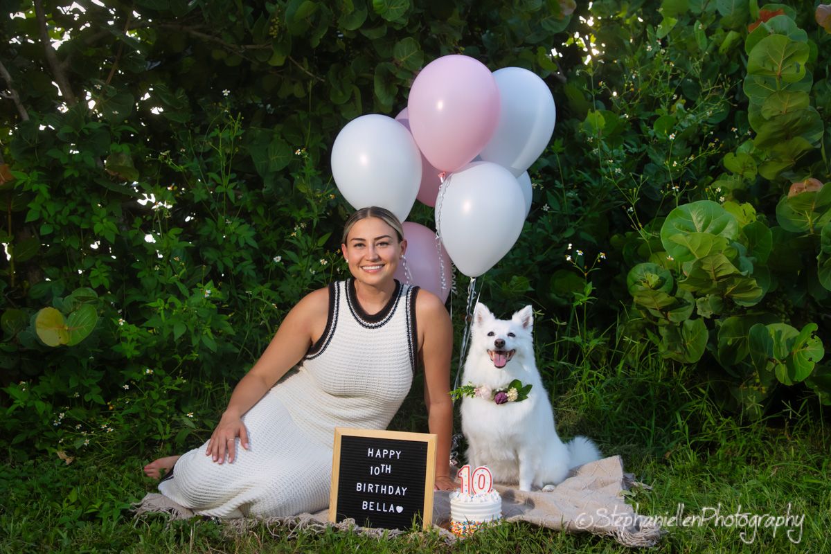 Woman sitting with white Samoyed dog beside birthday balloons and sign.