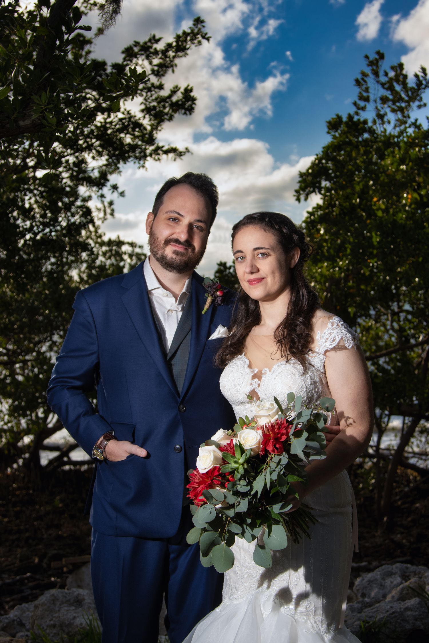Wedding couple poses outdoors, bride holding red and white bouquet, groom in navy suit under dramatic cloudy sky.