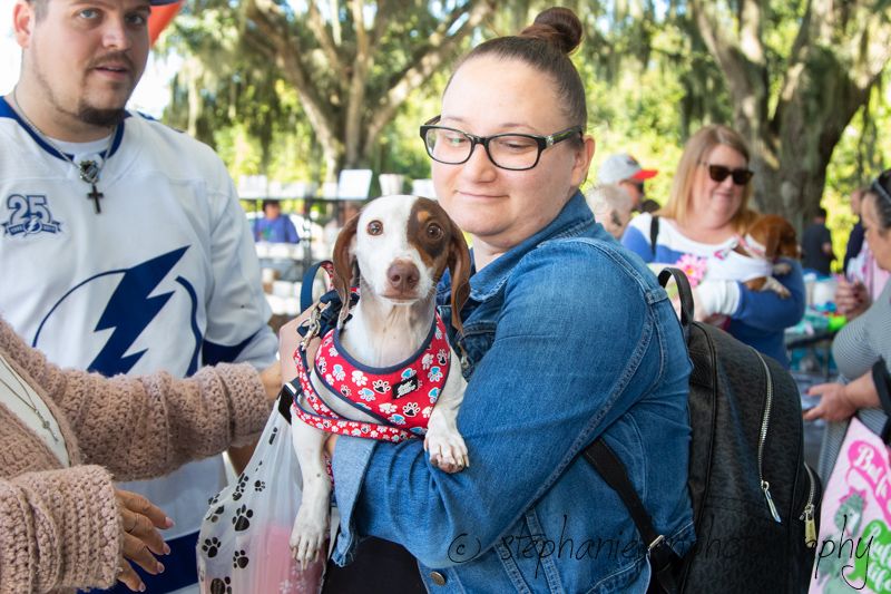 Woman holding a spotted dog wearing a red bandana at an outdoor pet event.
