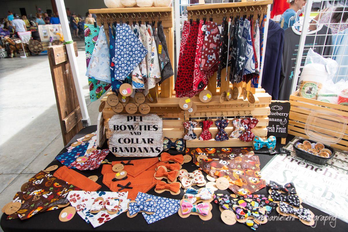 Vendor booth displaying colorful pet bow ties, collar bandanas, and accessories on wooden display stand.