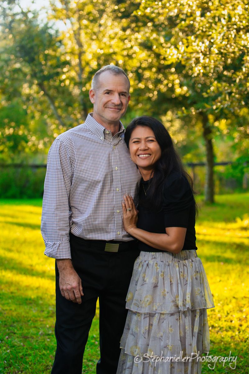 Smiling couple posing together outdoors in a sunny green park, man in checkered shirt and woman in black top and floral skirt.