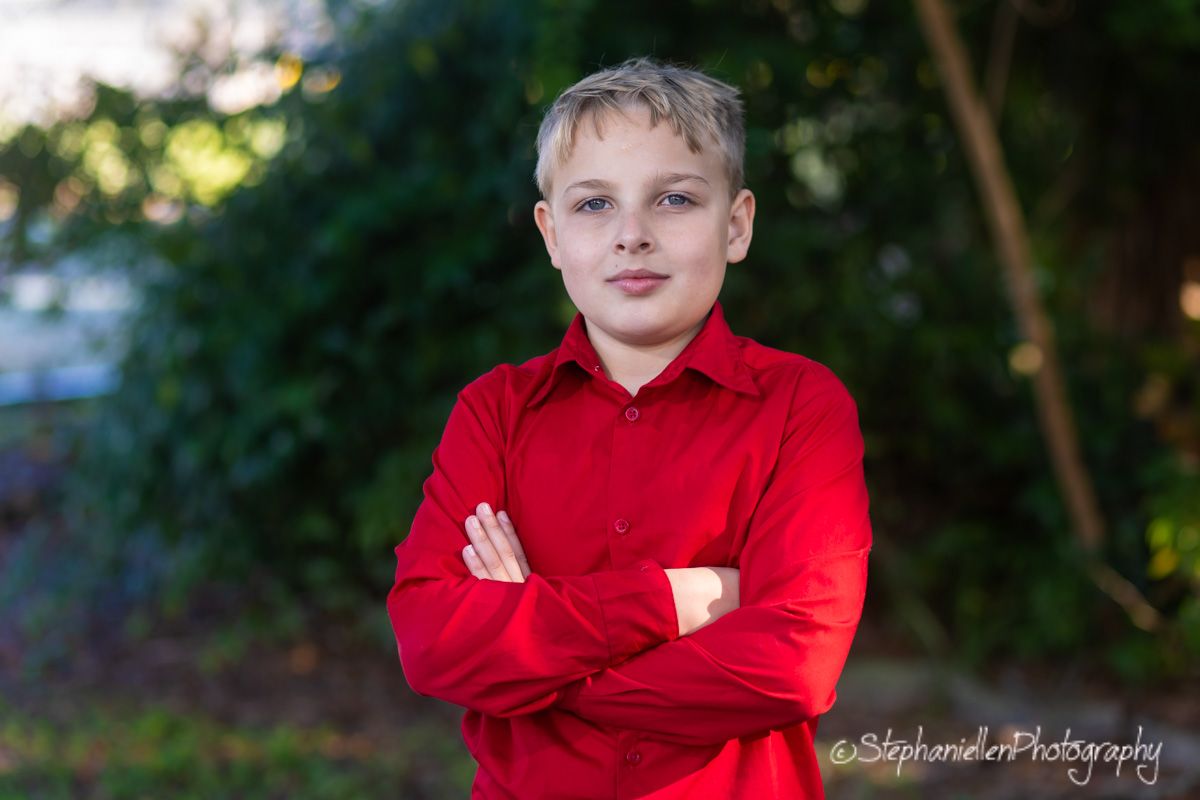 Young boy in red shirt standing with arms crossed outdoors, professional portrait photography.