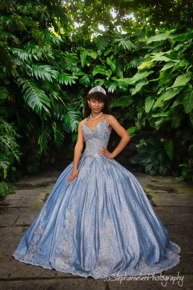 Young woman in blue ball gown with tiara standing in lush tropical garden.