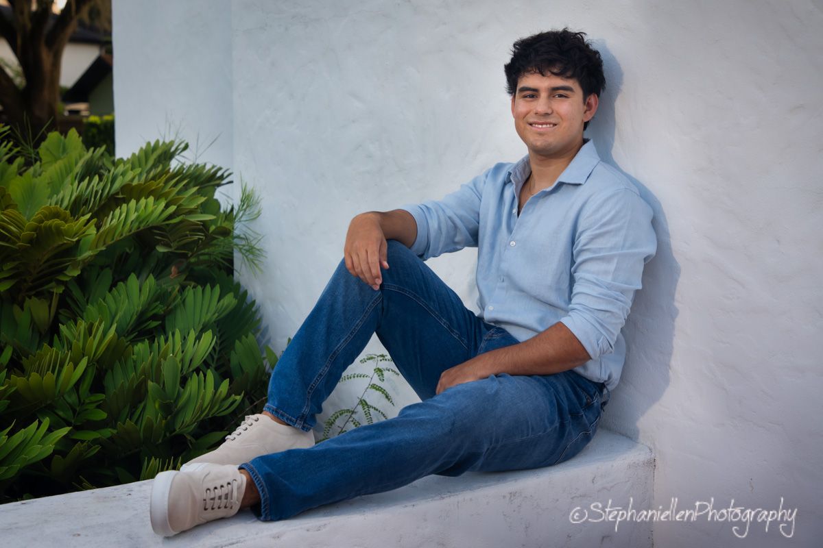 Young man in light blue shirt and jeans sitting against white wall with tropical greenery.