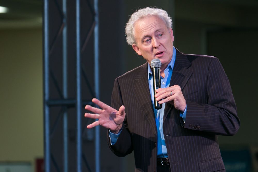 Older man in pinstripe suit speaks passionately into microphone at indoor event.