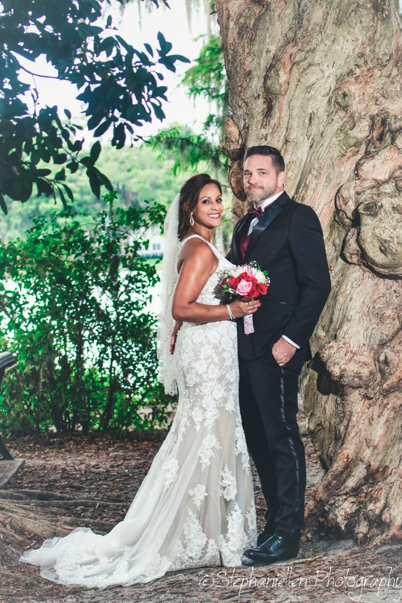 Bride in fitted lace gown and groom in black tuxedo pose together beside a massive tree near a waterfront.