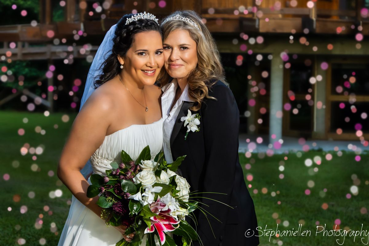 Two brides pose together with confetti falling, one in white gown with veil and bouquet, other in black suit.