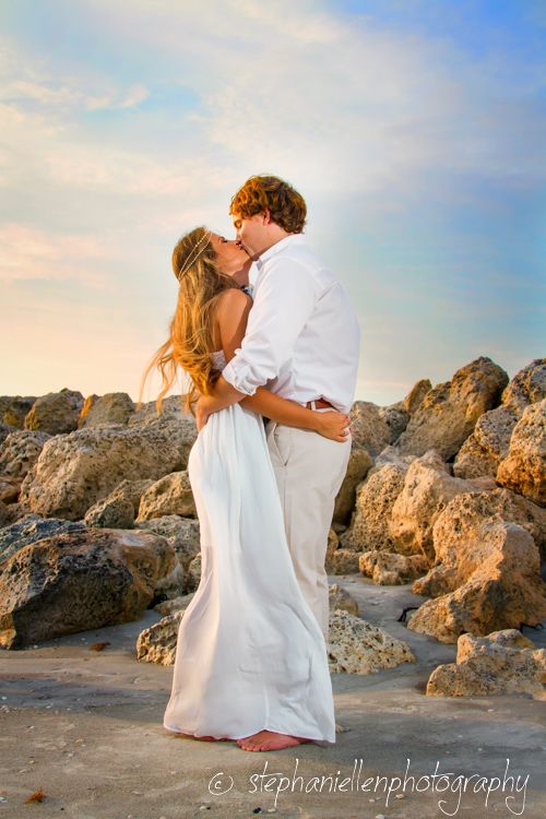 Couple kissing in white outfits on a rocky beach during a golden sunset.