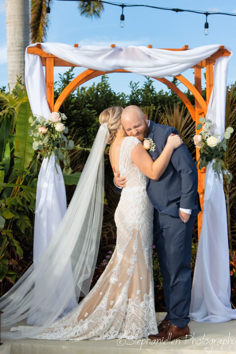 Bride and groom share their first kiss under a draped wooden arch adorned with white flowers at outdoor wedding.