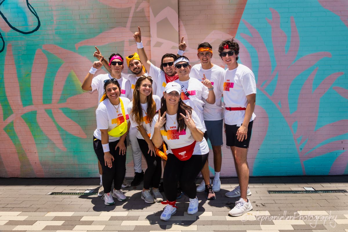 Group of young people in white shirts pose together in front of colorful tropical mural.
