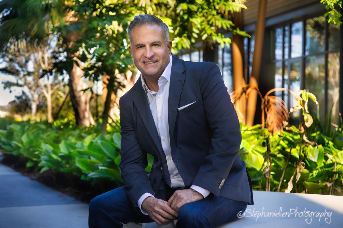 Professional headshot of a smiling man in navy suit seated outdoors among lush greenery.