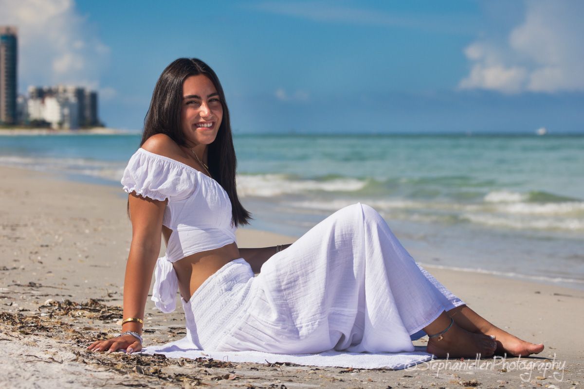 Young woman in white crop top and skirt sitting on sandy beach smiling on a sunny day.