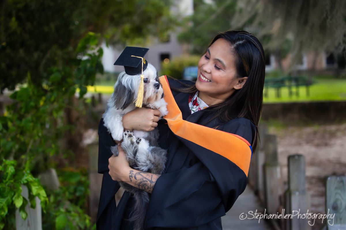 Smiling graduate in cap and gown holding a small dog wearing a graduation cap outdoors.