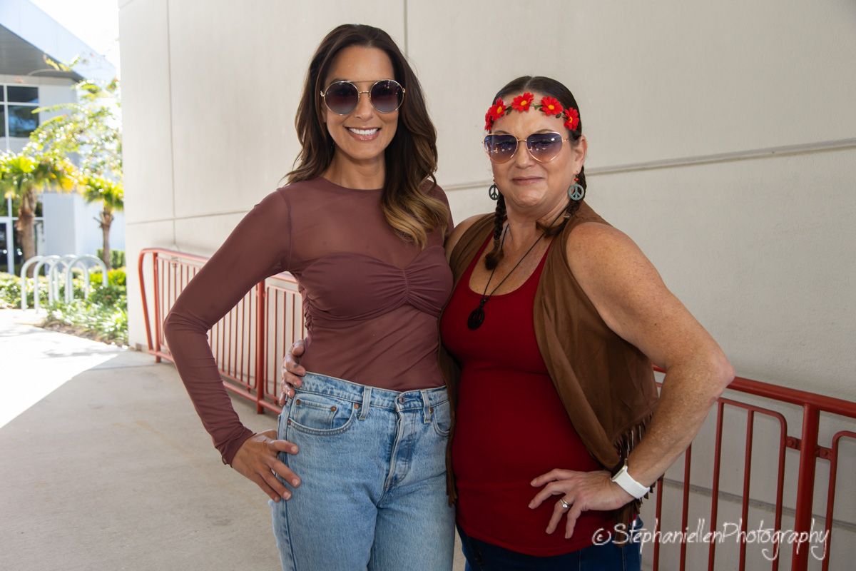 Two women wearing sunglasses posing together outdoors on a sunny day near a striped fence.
