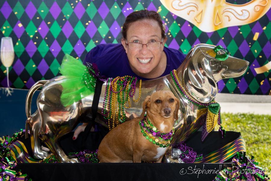 Man poses with dachshund dog wearing Mardi Gras beads beside decorative dachshund balloon at festive party.
