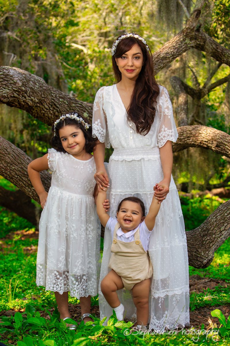 A mother in white lace dress poses with her two young children in a lush green park setting.