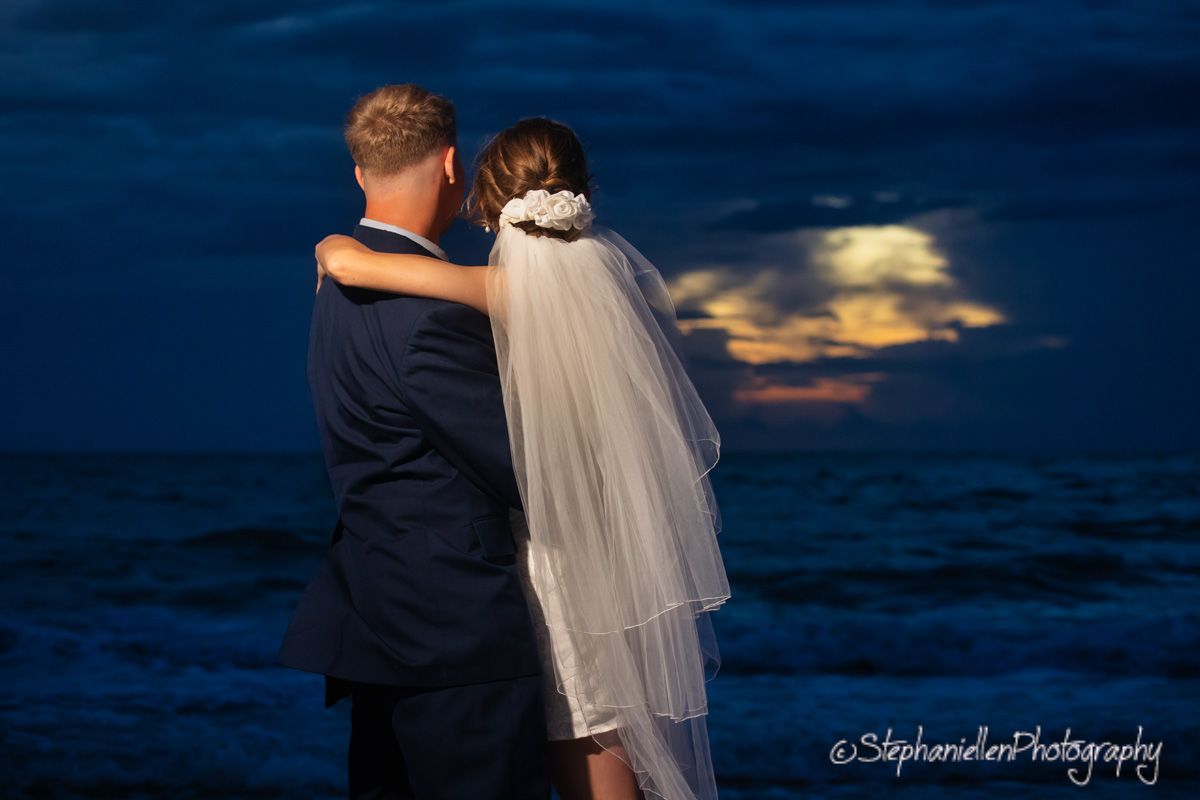 Bride and groom embrace on beach at night with full moon glowing over ocean waters.
