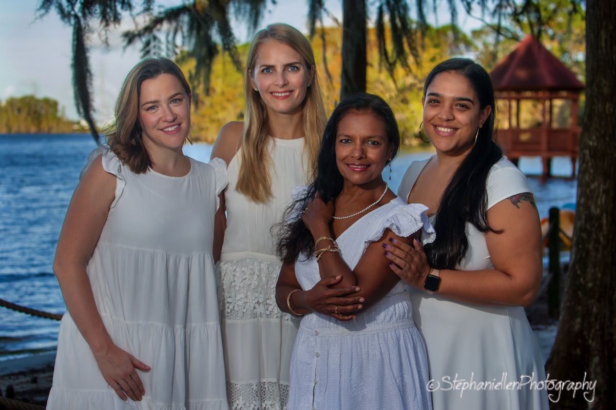 Four women in white dresses smile together outdoors near a lake with a gazebo at golden hour.