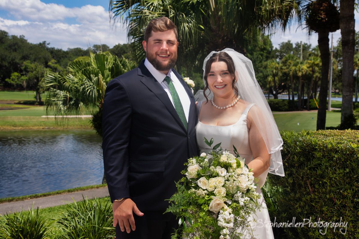 Smiling bride and groom posing outdoors near a pond with lush greenery on their wedding day.