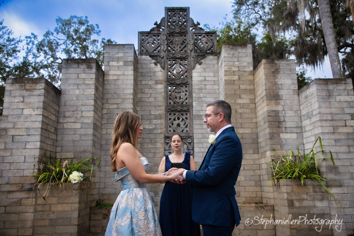 Couple exchanges vows before ornate stone cross at outdoor wedding ceremony with officiant.