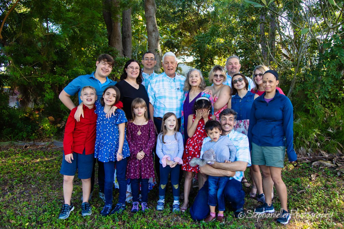 Large multigenerational family group portrait outdoors, smiling together among lush green trees in natural light.