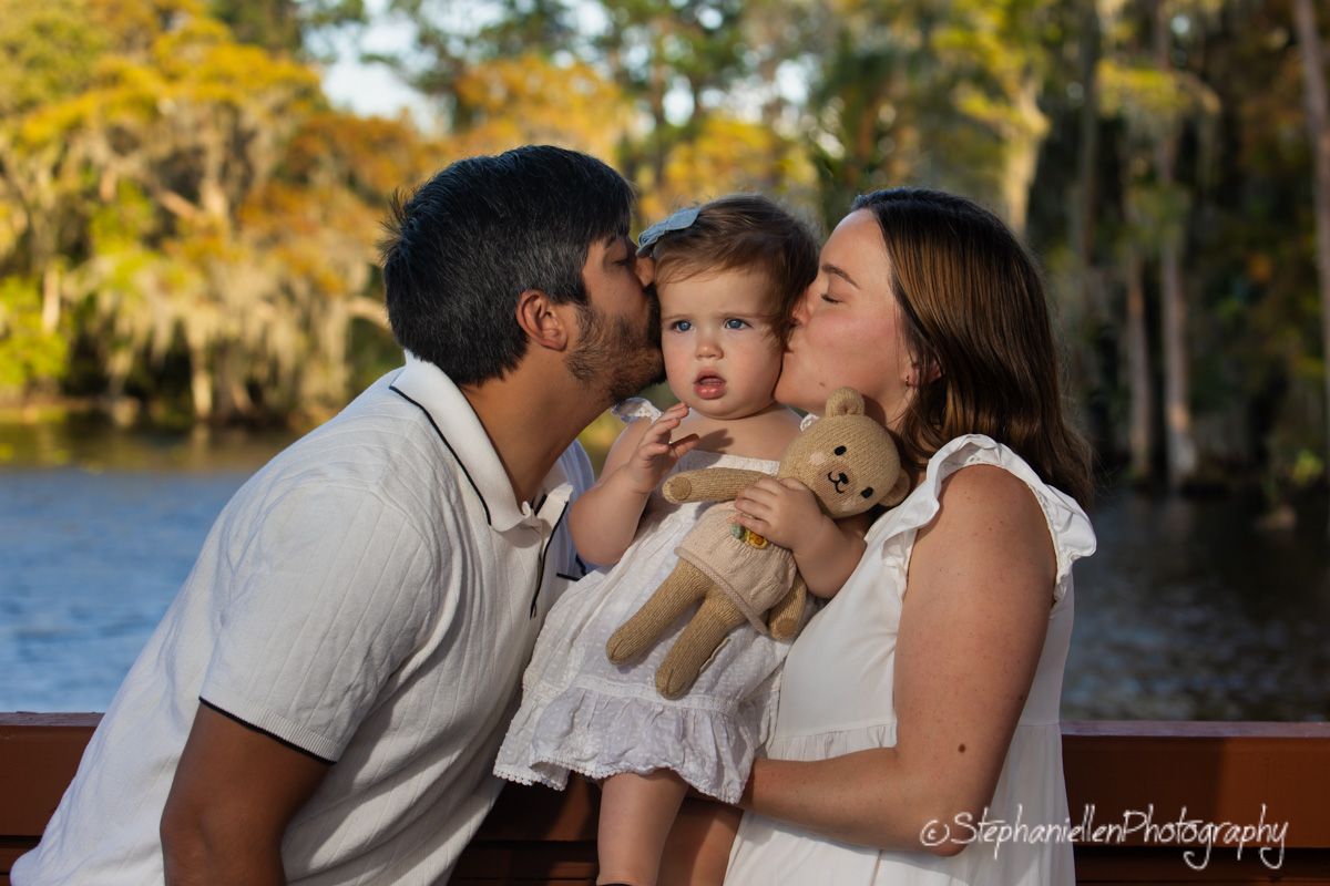A couple kisses their toddler girl holding a teddy bear outdoors near a scenic waterway with lush green trees.