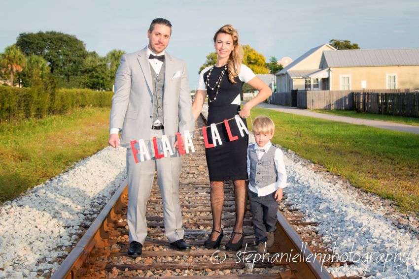 Family of three posing on railroad tracks, parents in formal attire holding banner, young boy in vest beside them.