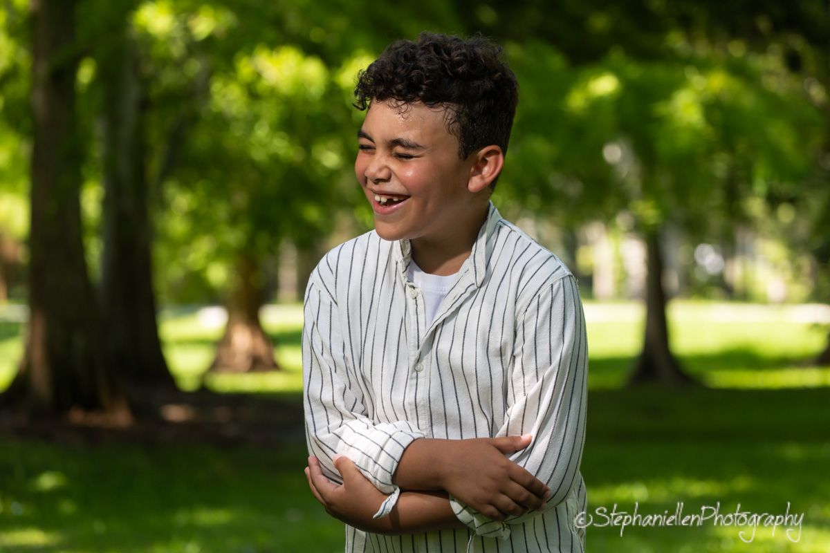 A laughing young boy with curly hair stands outdoors in a park, arms crossed, wearing a striped shirt.