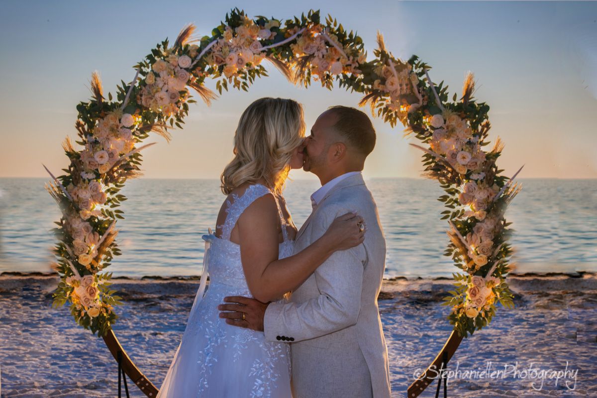 Couple sharing a kiss under a floral arch at a stunning beach wedding sunset.