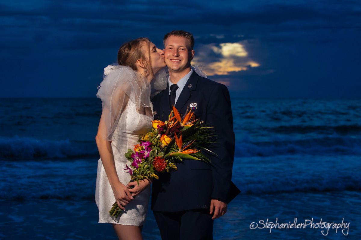 A bride kisses her grooms cheek at twilight on a beach, he holds a tropical bouquet with the ocean behind them.